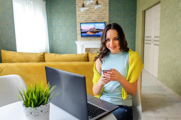 Foto hermosa joven moderna está sentada en la mesa con una computadora y un teléfono en las manos ropa elegante y brillante estilo juvenil ambiente acogedor en el hogar