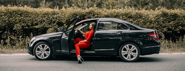 Una hermosa joven con un mono rojo se sienta al volante de un coche negro en una carretera vacía en el bosque