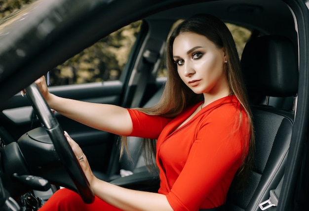 Una hermosa joven con un mono rojo se sienta al volante de un coche negro en una carretera vacía en el bosque