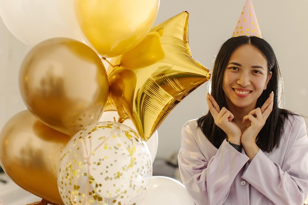 Hermosa joven morena asiática con globos mirando a la cámara cruzando los dedos y pidiendo deseos Conceptos de fiesta de cumpleaños