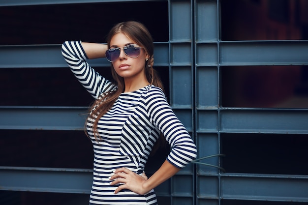 Hermosa joven morena con bonito vestido a rayas, gafas de sol. Posando sobre fondo urbano. Foto de moda