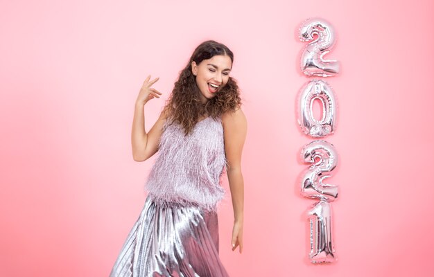 Hermosa joven morena con cabello rizado en ropa festiva se ve feliz en una pared rosa con globos plateados para el concepto de año nuevo