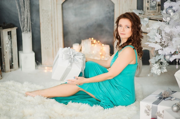 Hermosa joven morena en elegante vestido azul sentado junto al árbol de Navidad blanco y regalos. Foto de glamour de año nuevo