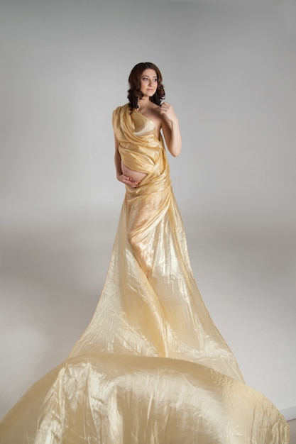 Hermosa joven morena embarazada con un paño amarillo transparente en foto de estudio aislado sobre fondo blanco. La modelo está de pie en alto abriendo su vientre.