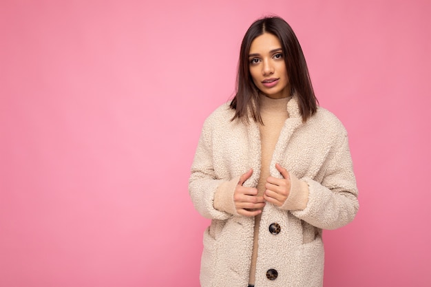 Hermosa joven morena europea vistiendo elegante abrigo cálido beaige aislado sobre fondo rosa con espacio de copia. Concepto de moda.