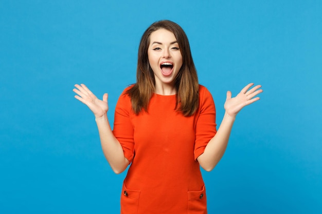 Hermosa joven morena feliz y emocionada con un vestido naranja rojo extendiendo las manos, diga sí aislada sobre un retrato de estudio de fondo de pared azul. Concepto de moda de estilo de vida de la gente. Simulacros de espacio de copia.