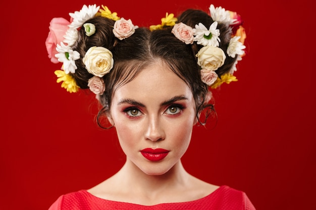 Hermosa joven morena con un increíble peinado floral con maquillaje posando aislada sobre pared roja, mirando al frente