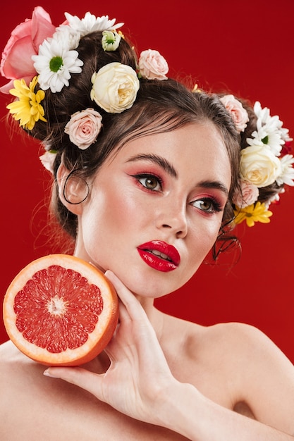 Hermosa joven morena con un increíble peinado floral con maquillaje posando con un pomelo aislado sobre pared roja