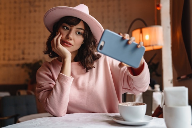 Una hermosa joven morena en el interior de la cafetería tomando café con teléfono móvil tomar un selfie.