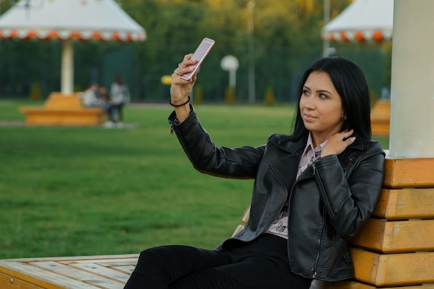 Hermosa joven morena con maquillaje y elegante manicura en chaqueta de cuero se sienta en un banco del parque y hace selfie