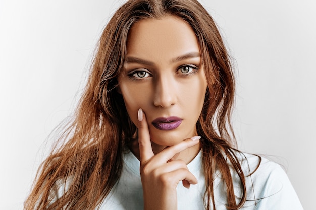 Hermosa joven morena de ojos verdes con cabello largo con maquillaje de moda con labios morados sobre un fondo gris aislado. La mujer se lleva la mano a la cara y mirando seriamente a la cámara.