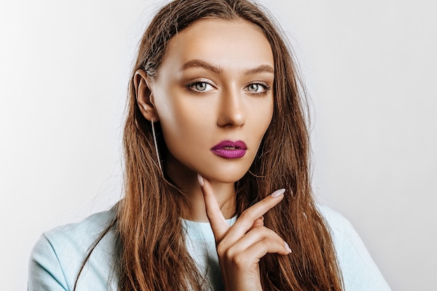 Hermosa joven morena con ojos verdes con cabello largo con maquillaje de moda con labios púrpuras en una pared gris aislada. La mujer se lleva la mano a la cara y mirando seriamente a la cámara