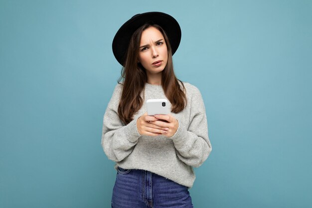 Hermosa joven morena pensando con sombrero negro y suéter gris con smartphone