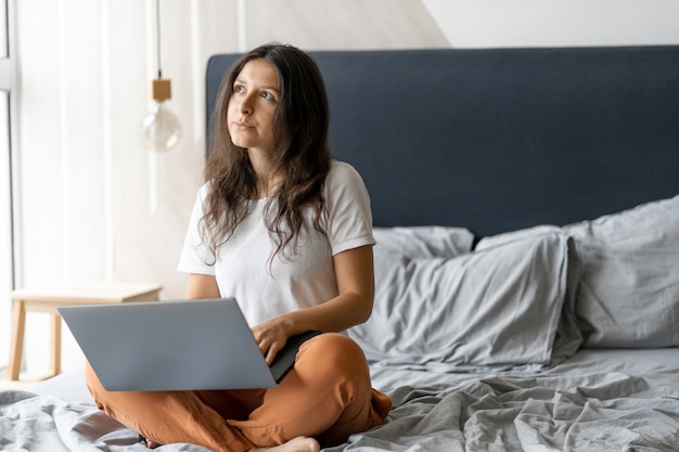 Hermosa joven morena con un portátil sentado en la cama. Elegante interior moderno. Un lugar de trabajo acogedor. Comprar en internet