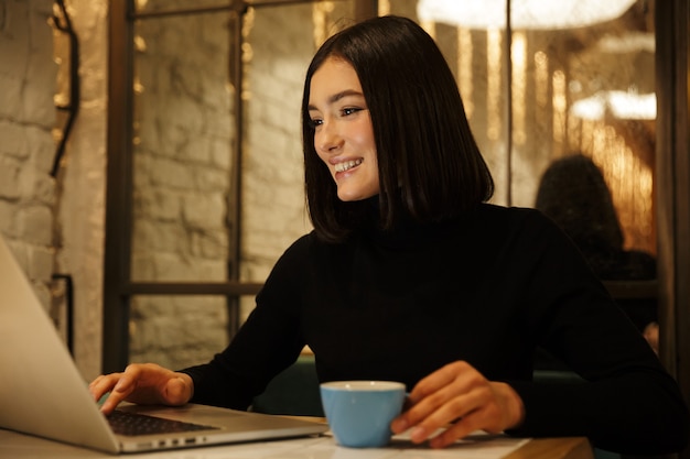 Hermosa joven morena sentada en la mesa de café en el interior, trabajando en equipo portátil