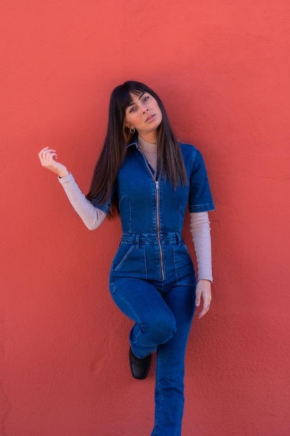 Hermosa joven morena sonriendo apoyada contra una pared con un traje de mezclilla azul. Bastante caucásica posando sobre un fondo rojo.