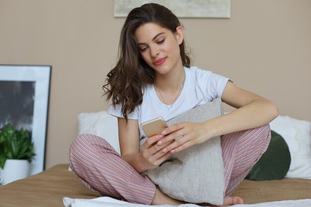 Hermosa joven morena sonriente acostada en la cama blanca y usando un teléfono en su dormitorio.