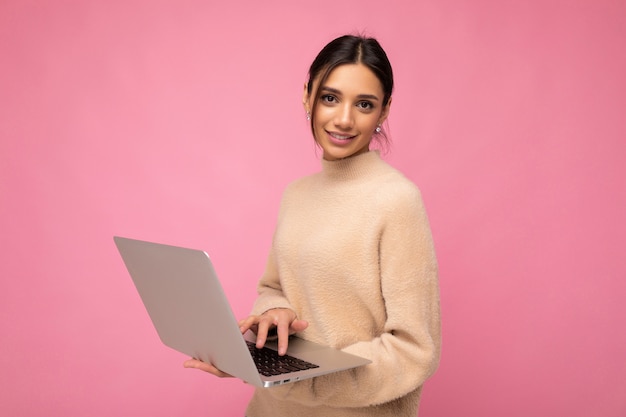 Hermosa joven morena vestida con un suéter beige sosteniendo la computadora netbook escribiendo texto en el teclado aislado sobre la pared rosa.