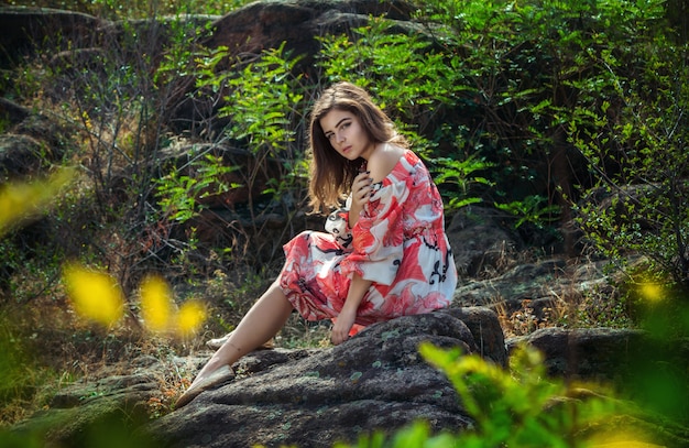 Hermosa joven morena con vestido colorido, sentado en un acantilado de piedra en un bosque en un caluroso día de verano