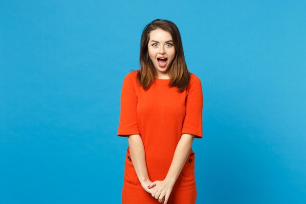 Hermosa joven morena con un vestido rojo anaranjado mirando una cámara de pie posando aislada sobre un moderno fondo de pared azul, retrato de estudio. Concepto de moda de estilo de vida de la gente. Simulacros de espacio de copia.