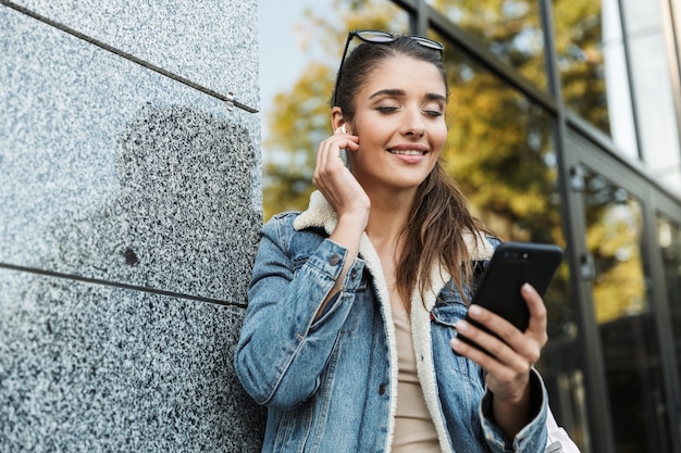 Hermosa joven morena vistiendo chaqueta, llevando mochila caminando al aire libre, mediante teléfono móvil