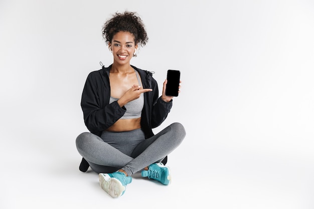 Hermosa joven mujer africana de fitness deportes asombrosos mediante teléfono móvil escuchando música que muestra la pantalla.