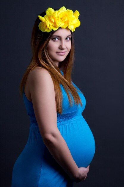 Hermosa joven mujer embarazada en vestido azul con flores amarillas en el pelo sobre fondo gris