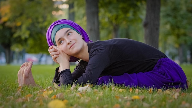 Hermosa joven mujer islámica usa hiyab sentada en la hierba en el parque al aire libre agachándose en el cuerpo estirando el vientre haciendo ejercicio de yoga entrenamiento físico descansando relajante sonriendo