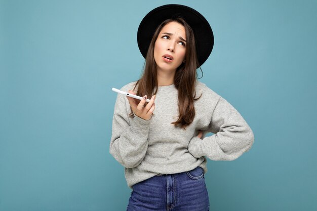 Hermosa joven mujer morena pensando con sombrero negro y suéter gris sosteniendo smartphone mirando mensajes de texto aislados en el fondo. Copiar el espacio