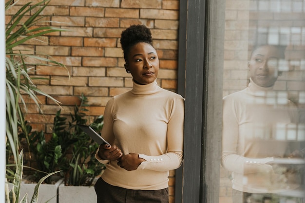 Una hermosa joven mujer de negocios afroamericana con tableta digital de pie junto a la pared de ladrillo en la oficina de estilo industrial