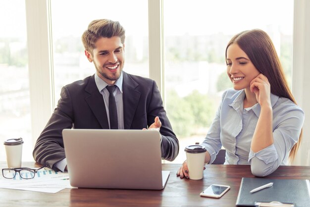 Hermosa joven mujer de negocios y apuesto hombre de negocios con trajes formales están usando una laptop hablando y sonriendo mientras trabajan en la oficina