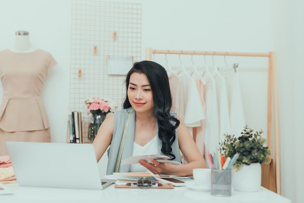 Hermosa joven mujer de negocios asiática / diseñadora de moda con computadora en el taller