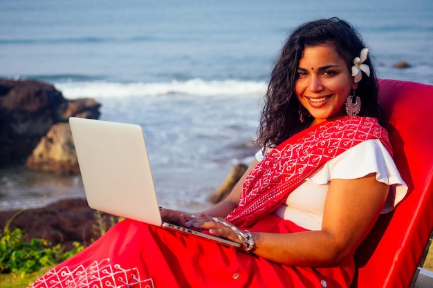 Hermosa y joven mujer de negocios india en el tradicional sari de la india trabajando con una computadora portátil de la estudiante de mar asiático trabajo remoto independiente sentado en la playa usando un teléfono inteligente