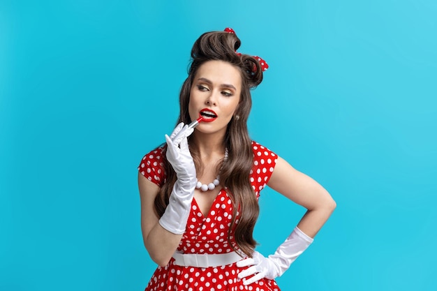 Hermosa joven mujer pinup en vestido retro aplicando lápiz labial rojo brillante sobre fondo de estudio azul