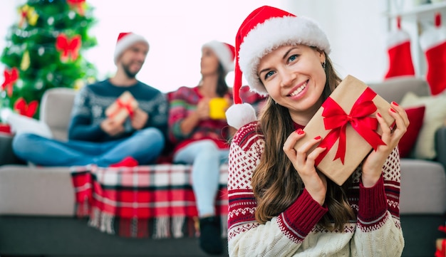 Hermosa joven mujer sonriente feliz en un suéter de Navidad y gorro de Papá Noel está sosteniendo una caja de regalo