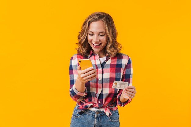Hermosa joven mujer vistiendo camisa a cuadros sosteniendo teléfono celular y tarjeta de crédito aislado