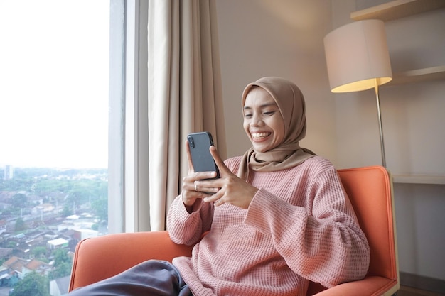 hermosa joven musulmana asiática usando hiyab mientras usa un teléfono inteligente sentado cerca de la ventana