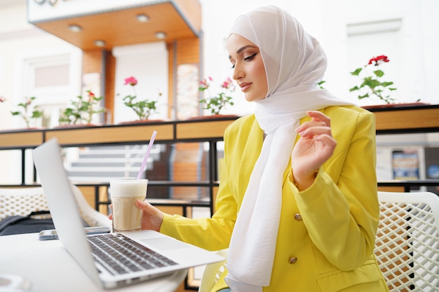 Hermosa joven musulmana usando laptop mientras está sentado en la cafetería