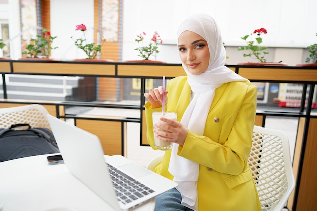 Hermosa joven musulmana usando laptop mientras está sentado en la cafetería