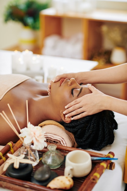 Hermosa joven negra disfrutando de un masaje facial y de cabeza en el salón de spa cuando está acostado en la cama junto a la bandeja con aceites aromáticos