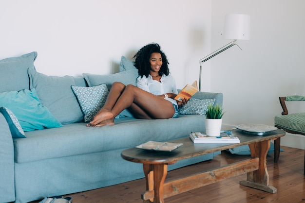 Hermosa joven negra sentada en el sofá leyendo un libro