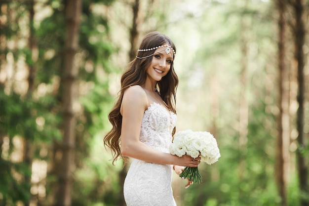 Hermosa joven novia con peinado de novia con joyas en vestido de encaje con un ramo de flores en sus manos y posando en el bosque temprano en la mañana