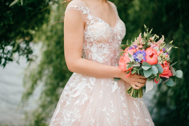 Hermosa joven novia en vestido de novia blanco posando al aire libre con ramo de flores