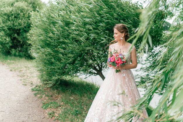 Hermosa joven novia en vestido de novia blanco posando al aire libre con ramo de flores