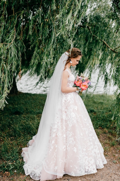 Hermosa joven novia en vestido de novia blanco posando al aire libre con ramo de flores