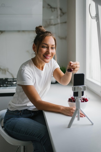 Hermosa joven nutricionista realiza una videollamada en línea a través de un teléfono inteligente con un amigo para hablar sobre los beneficios de las cerezas frescas en la cocina de casa