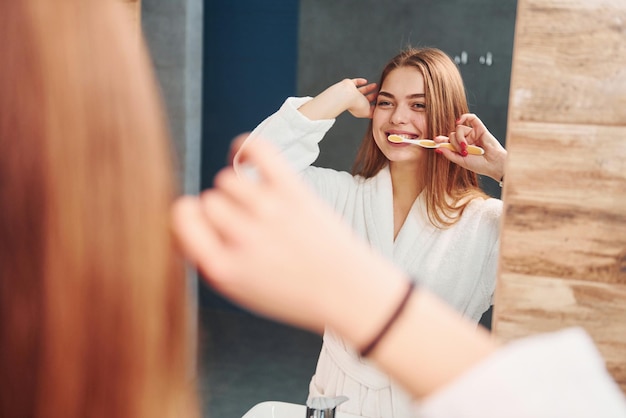 Hermosa joven parada en el baño cerca del espejo y cepillándose los dientes