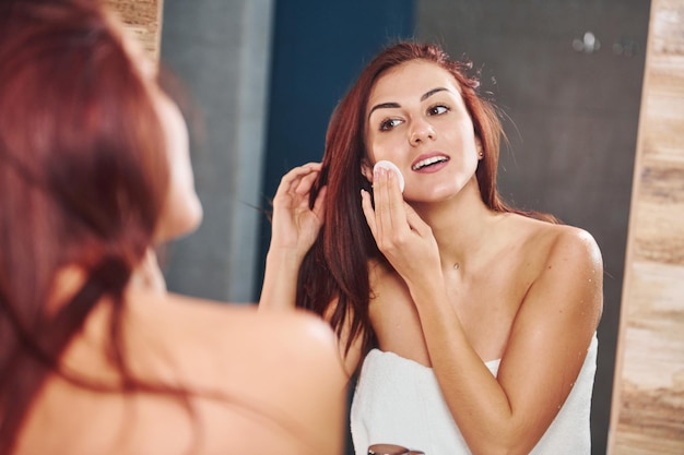 Hermosa joven parada en el baño, mirándose al espejo y cuidando su rostro.