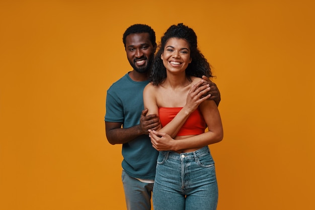 Hermosa joven pareja africana sonriendo y abrazándose mientras está de pie contra el fondo amarillo