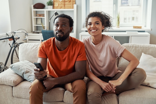 Hermosa joven pareja africana viendo la televisión y sonriendo mientras pasa tiempo en casa